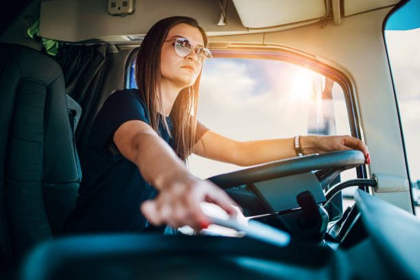 Portrait of beautiful young woman professional truck driver sitting and driving big truck. She is dangerously trying to take smart phone while driving.